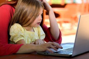 Przy otwartym laptopie siedzi kobieta w czerwonej bluzce. Na jej kolanach siedzi dziewczynka w żółtej sukience i wpatruje się w komputer.
