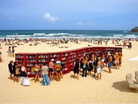 Regały z książkami na plaży w Australii. Wokół stoi tłum ludzi