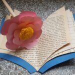Książeczka wykonana z kartek starej książki, z niebieską okładką. na książce leży różowy kwiat z papieru, z żółtym środkiem z krepiny