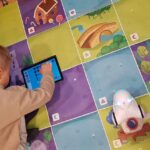 Na dywanie leży mata do kodowania. Przedszkolak rysuje na tablecie drogę robota. Obok stoi robot Photon