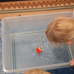 Dziewczynka dmucha na mały stateczek z korka, ustawiony na wodzie w plastikowym pojemniku