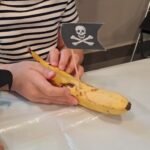 Dziecko trzyma w ręce banana z piracką flagą. Obok stoi piracka papuga z rolki po papierze toaletowym, przyozdobiona piórkami kreatywnymi