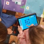 Przedszkolaki siedzą obok maty do kodowania. Dziewczynka rysuje na tablecie drogę robota