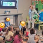 Przedszkolaki siedzą na dywanie i słuchają bibliotekarki, która trzyma w ręce smoka wykonanego z materiałów do recyklingu