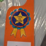 Pomarańczowa zawieszka na drzwi z kotylionem, w środku którego jest gwiazda z napisem Tata na medal. Poniżej napis: Tata odpoczywa!