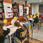 Uczniowie siedzą przy stolikach w bibliotece i kolorują ilustracje. W tle regały z książkami i